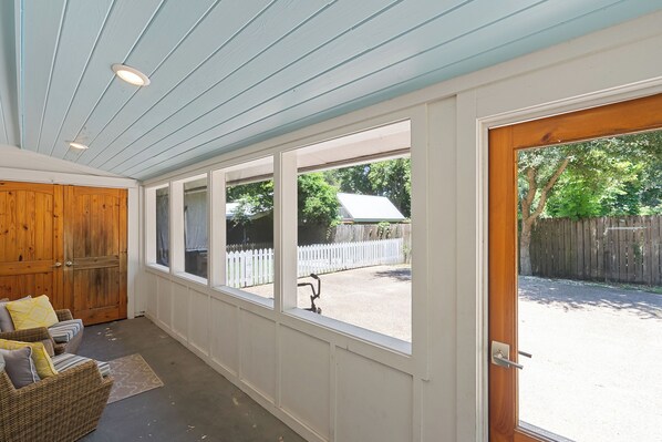 A covered porch with a view of a backyard, a wooden door, and a wicker sofa with cushions.