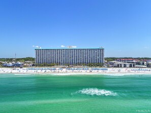 A large hotel with multiple floors and balconies, situated on a beach with clear turquoise waters.
