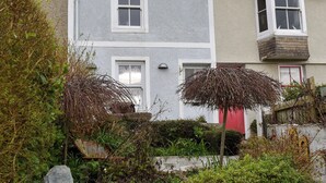 A two-story house with a grey and white exterior, a red door, and a small balcony with a wooden railing. - 2 bedroom accommodation in St Ives (St Ives)
