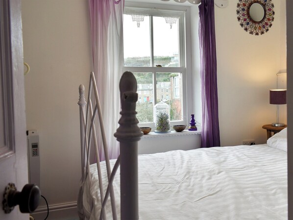 A bedroom with a bed, a window with curtains, a bedside table with a lamp, and a decorative mirror on the wall.