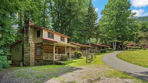 Une rangée de chalets aux cheminées en pierre et aux toits rouges, entourés d’une végétation luxuriante et bordés d’une allée de gravier.