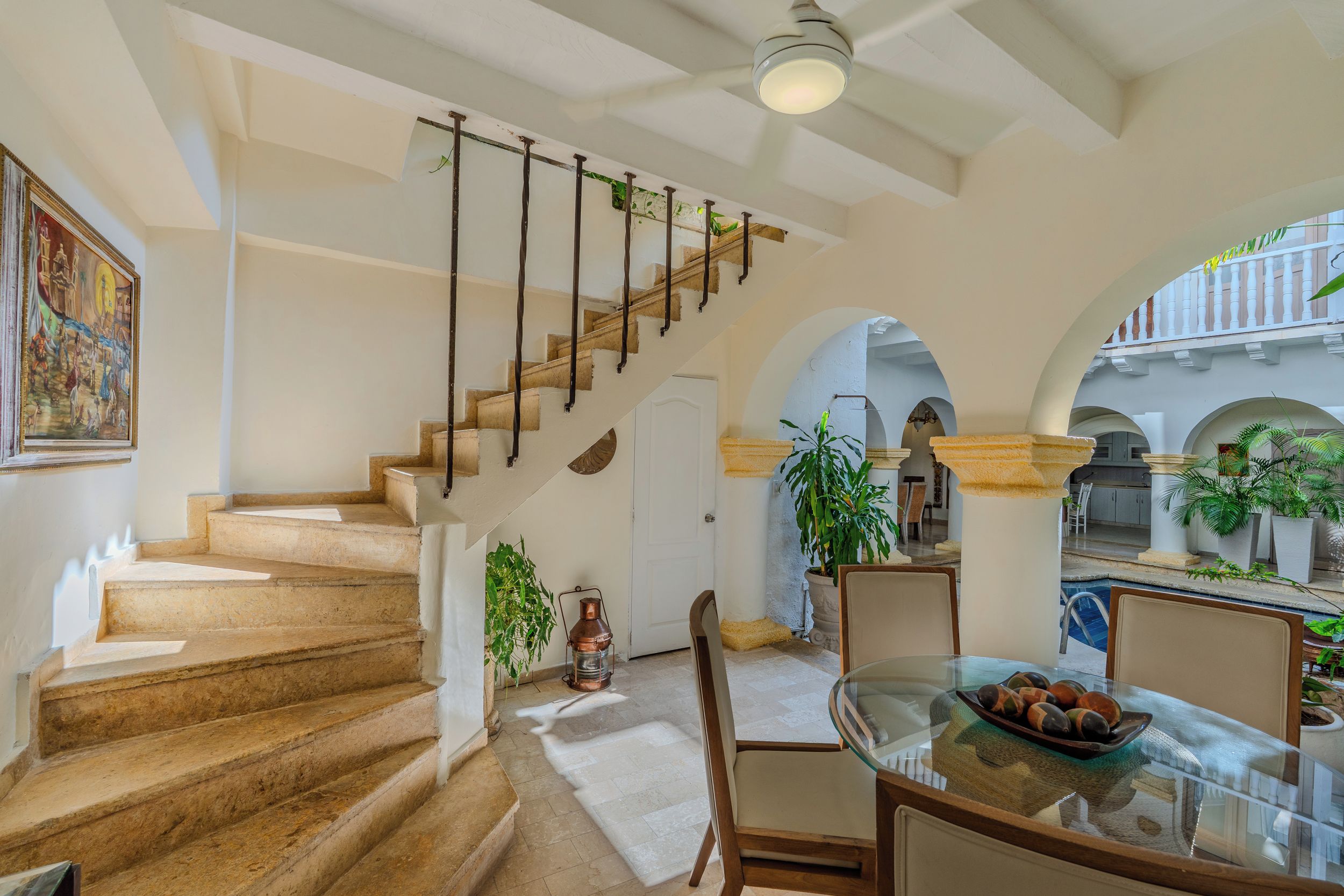 A staircase with a wooden handrail leading to a door, a dining area with a glass table and chairs, and a view of an outdoor courtyard with arches and plants.
