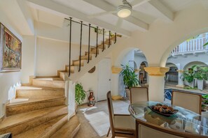 Un escalier avec une rampe en bois menant à une porte, un coin repas avec une table et des chaises en verre, et une vue sur une cour extérieure agrémentée d’arcades et de plantes.