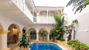 Una piscina con suelo de baldosas, rodeada de columnas, plantas en macetas y una escalera que lleva a un nivel superior.
