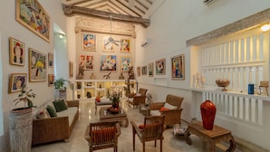Amplia sala de estar con techo alto, vigas de madera a la vista y una variedad de obras de arte en las paredes. Varias áreas de descanso con muebles de mimbre y un gran jarrón rojo sobre una mesa de madera.