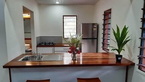 A compact kitchen with a central island, two stools, a refrigerator, and a window. - Whispering Palms Deluxe Villa - Absolute Beachfront (Siviri)