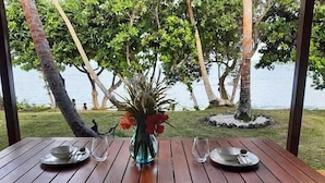 A wooden table with two plates and a vase of flowers, set against a backdrop of trees and a body of water. - Whispering Palms Deluxe Villa - Absolute Beachfront (Siviri)