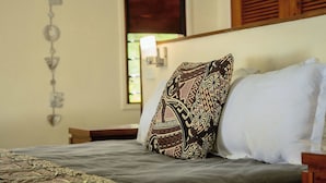 A bed with patterned and plain pillows, a decorative wall hanging, and a window with natural light. - Whispering Palms Deluxe Villa - Absolute Beachfront (Siviri)