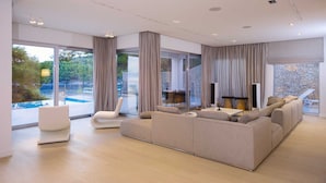 Un salón amplio con un sofá beige grande, un sillón blanco y una mesita de centro. Hay una pared de piedra y un ventanal con vistas a una piscina.