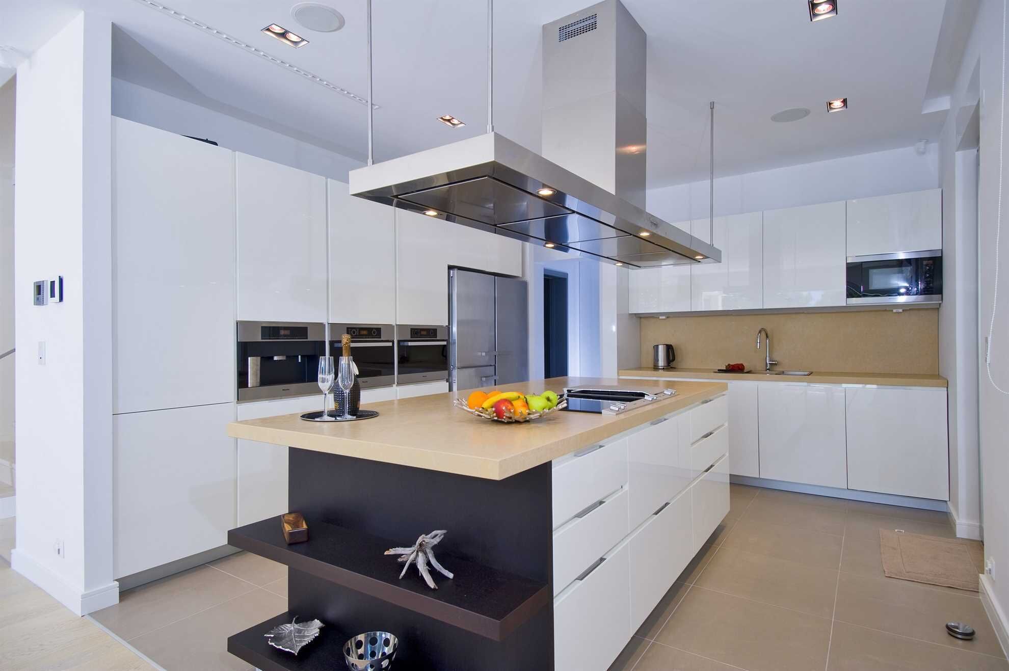 A modern kitchen with white cabinetry, a central island with a wooden countertop, built-in ovens, a stainless steel range hood, and a microwave.