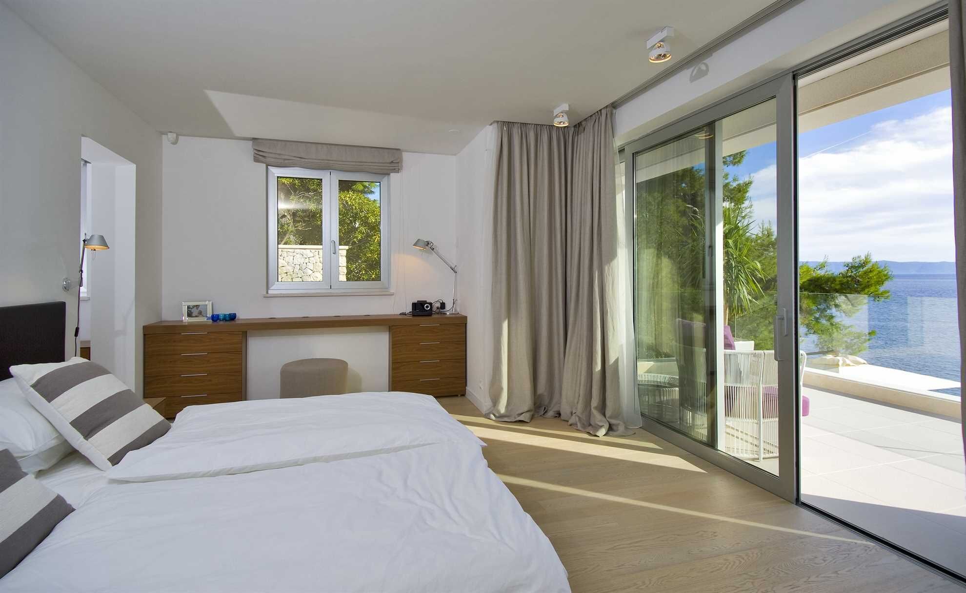A bedroom with a bed, a desk, a chair, and a view of the sea through large windows.