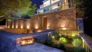 Un edificio moderno con fachada de piedra, un balcón y un espacio exterior bien iluminado con plantas y una zona de estar.