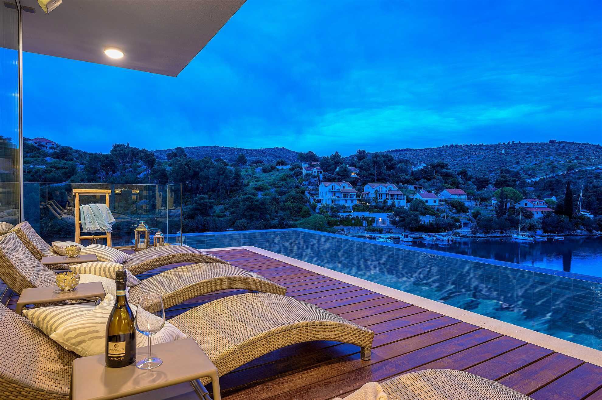 A balcony with wicker furniture and a bottle of wine, overlooking a body of water and houses.