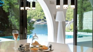 A table with a glass of wine, a tray of food, and a view of a pool and greenery through large windows. - Luxury Villa Notturno with Infinity Pool (Selca)