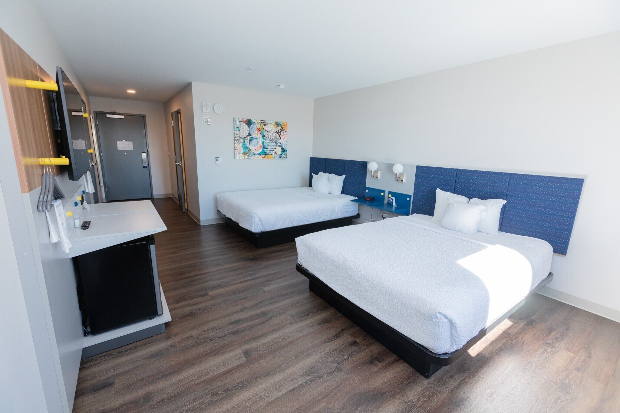 A hotel room with two beds, a desk, a mirror, a small cabinet, and a wall-mounted artwork.