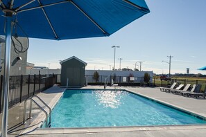 Una piscina con una sombrilla azul, un edificio pequeño y un área cercada con sillones de descanso.