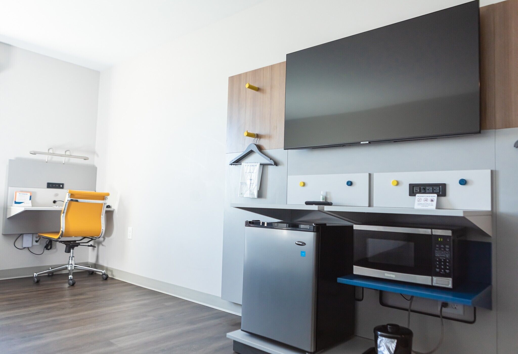Un espacio de oficina moderno con televisor de pantalla plana, refrigerador, microondas, escritorio con silla y un estante pequeño con un objeto colgante.