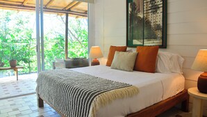 A bedroom with a large bed, a nightstand, a lamp, a framed picture on the wall, and a view of greenery outside. - In the gated community of Mar-A-Mar, walking distance from La Lora famous surf break and nicest white sand beach, the home counts with 3 bedrooms, 2 bathrooms, nice living room with open space kitchen & terraces. 24/7 security and shared pool. (Playa Santa Teresa de Cobano)