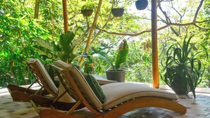 A lounge chair with a cushion, positioned on a tiled floor, surrounded by potted plants and a bamboo structure. - In the gated community of Mar-A-Mar, walking distance from La Lora famous surf break and nicest white sand beach, the home counts with 3 bedrooms, 2 bathrooms, nice living room with open space kitchen & terraces. 24/7 security and shared pool. (Playa Santa Teresa de Cobano)