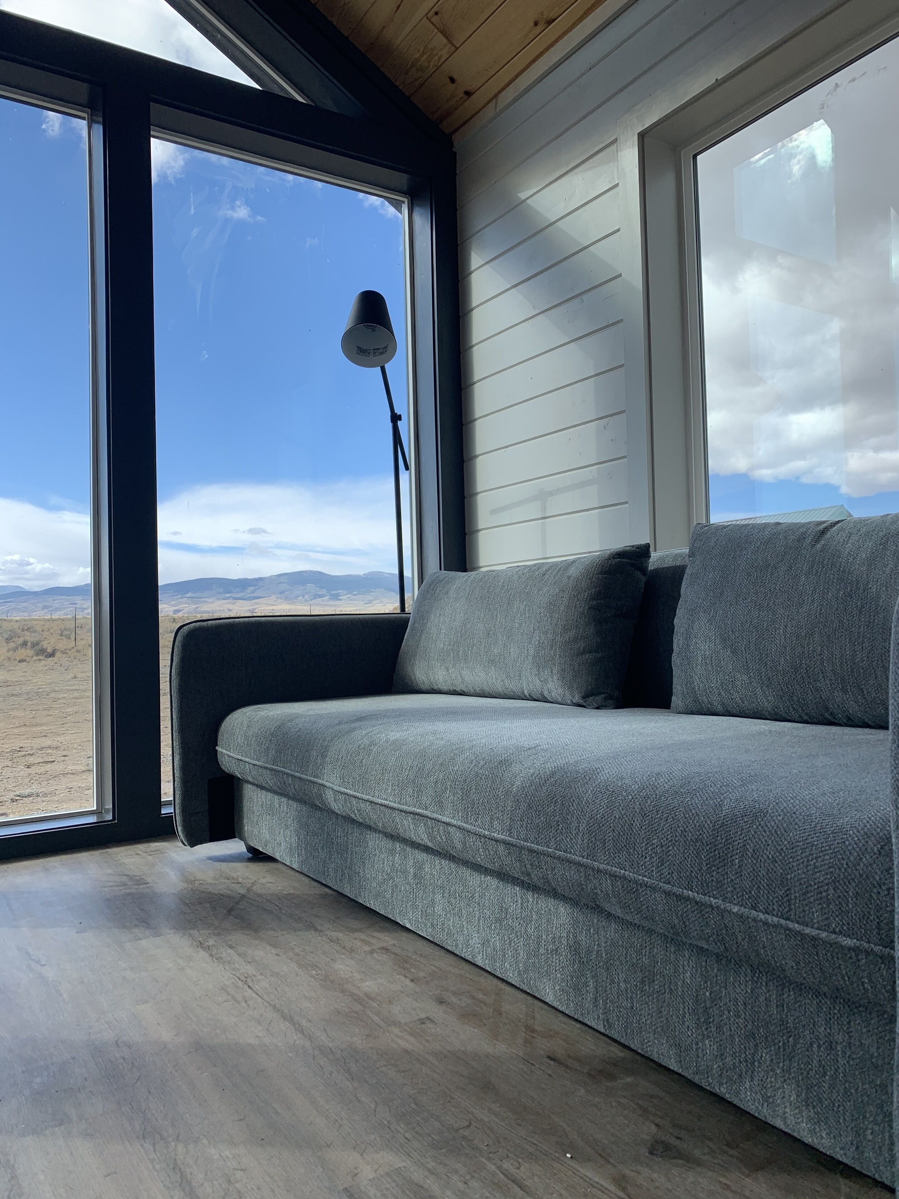 Ein Zimmer mit grauem Sofa, großen Fenstern und einer Holzboden.