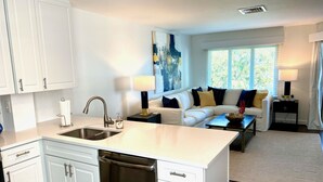 A modern kitchen with white cabinetry and a dishwasher, a living area with a sofa and a coffee table, and a window with a view of greenery. - Mid Century Modern meets Beach Chic, Beach views/ pool access,302G Port O Call (Isle of Palms)