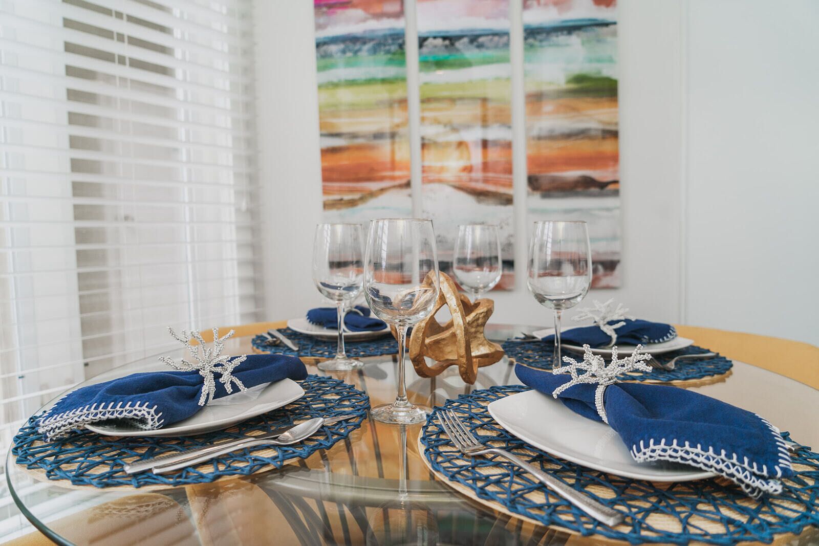 Una mesa de comedor dispuesta con servilletas azules, cubiertos y copas de vino, con un centro de mesa dorado decorativo.