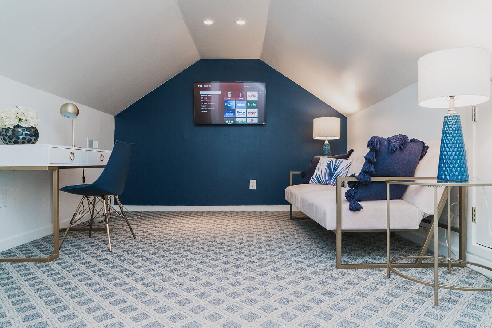 Una habitación moderna con una pared de acento azul oscuro, un televisor montado, un escritorio con una silla, una cama con ropa de cama azul y un suelo estampado.