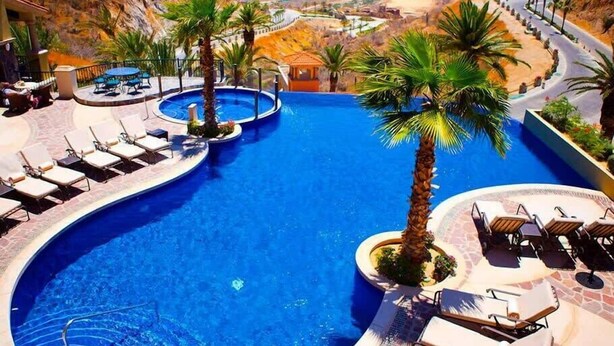 A large, curved swimming pool with multiple lanes, surrounded by lounge chairs and palm trees, set against a mountainous backdrop. - Gorgeous 3BR Villa Perfect View at Cabo San Lucas (Cabo San Lucas)