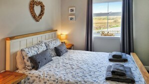 A neatly made bed with a patterned duvet, pillows, and folded towels. There is a bedside table with a lamp and a wall-mounted picture frame. A window with curtains is visible, offering a view of the outdoors. - 2 bedroom accommodation in Tayvallich (Tayvallich)
