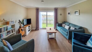 A living room with a sofa, a coffee table, a bookshelf, a guitar, and a television. - 2 bedroom accommodation in Tayvallich (Tayvallich)