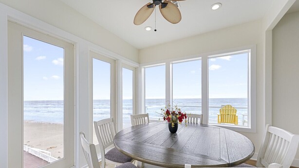 Una habitación con una mesa redonda grande, sillas blancas, un ventilador de techo y ventanas amplias con vista al mar.