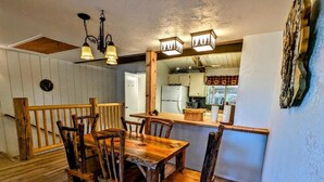 A rustic dining area with a wooden table and chairs, a kitchen in the background, and a decorative wall piece. - Your All-Season Chalet Basecamp – A/C + Unmatched Location! (Big Bear Lake)