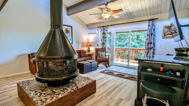 A cozy living room with a wood-burning stove, a ceiling fan, and a view of the outdoors through large windows. - Your All-Season Chalet Basecamp – A/C + Unmatched Location! (Big Bear Lake)
