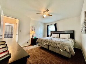 Un dormitorio con una cama, una mesita de noche, una lámpara, un ventilador de techo y una puerta que da a otra habitación.
