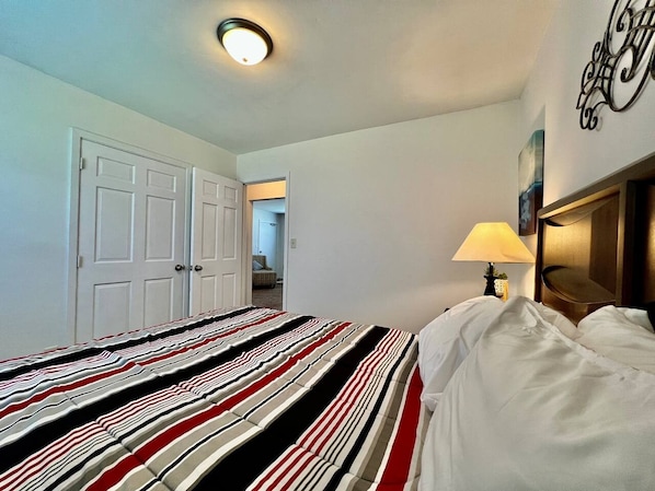 Een hotelkamer met een bed, nachtkastje, lamp, deur en een gang naar een andere kamer.