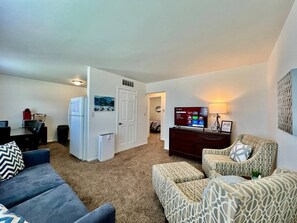 Sala de estar con sofá, otomanos y sillones estampados. Se ve un televisor, un refrigerador pequeño y un comedor.