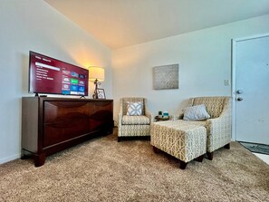 Una sala de estar con un televisor de pantalla plana, un mueble de madera, dos sillones estampados, una mesita y una puerta.