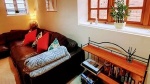 Un salón acogedor con un sofá de cuero marrón oscuro, una pequeña mesita de madera y una estantería llena de libros. Se ve una ventana con vistas a un edificio de ladrillo.
