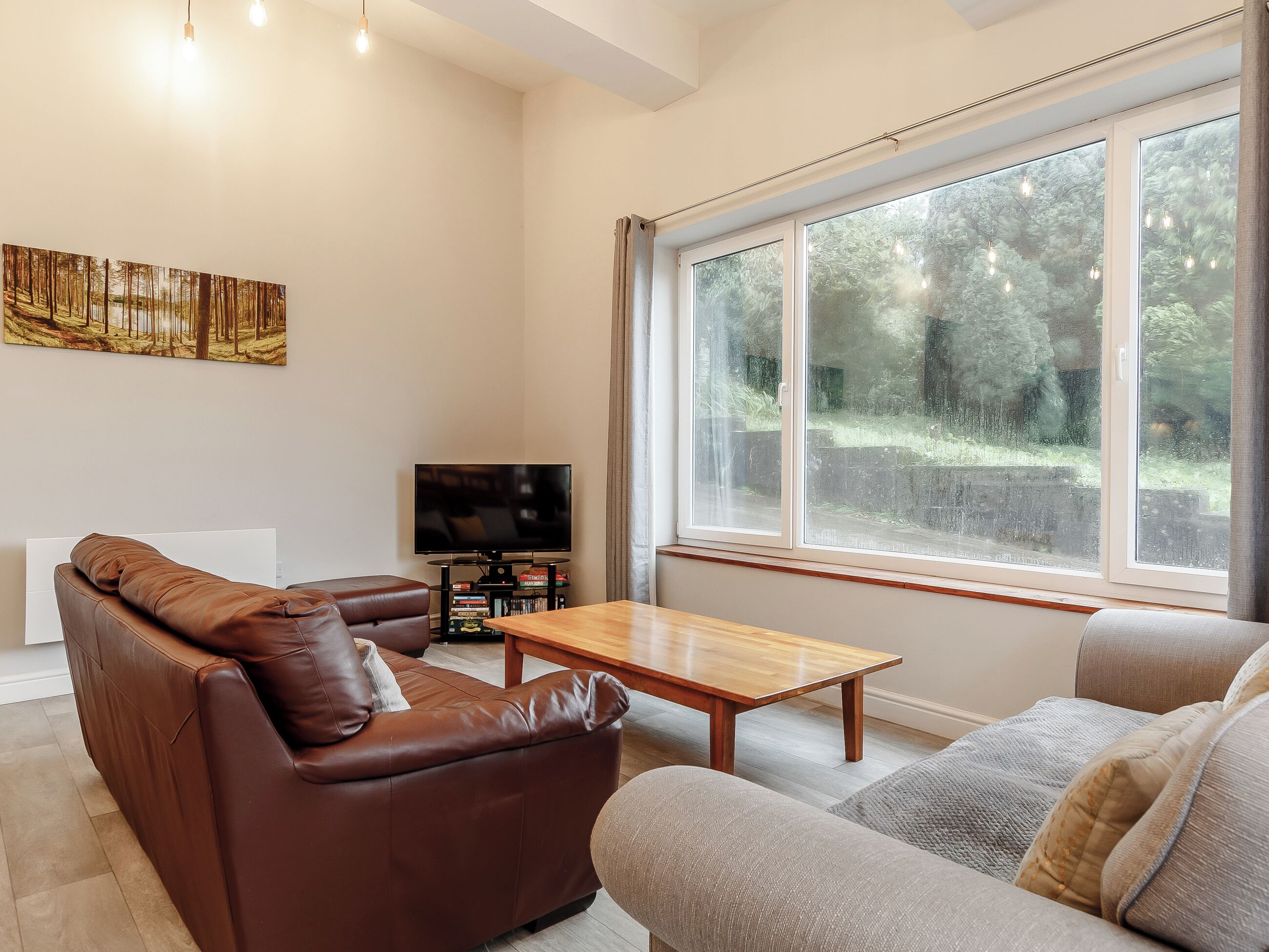 A living room with a brown leather sofa, a grey sofa, a wooden coffee table, a flat-screen TV, and a large window with a view of trees.