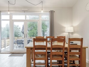 Et spiseområde med et træbord og stole, en lampe samt store vinduer med udsigt til det fri.