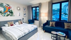 A hotel room with a bed, a sofa, a small table, and a view of the city through large windows. - New Modern Studio - Artur, Sveti Vlas - Apartello (Sveti Vlas)