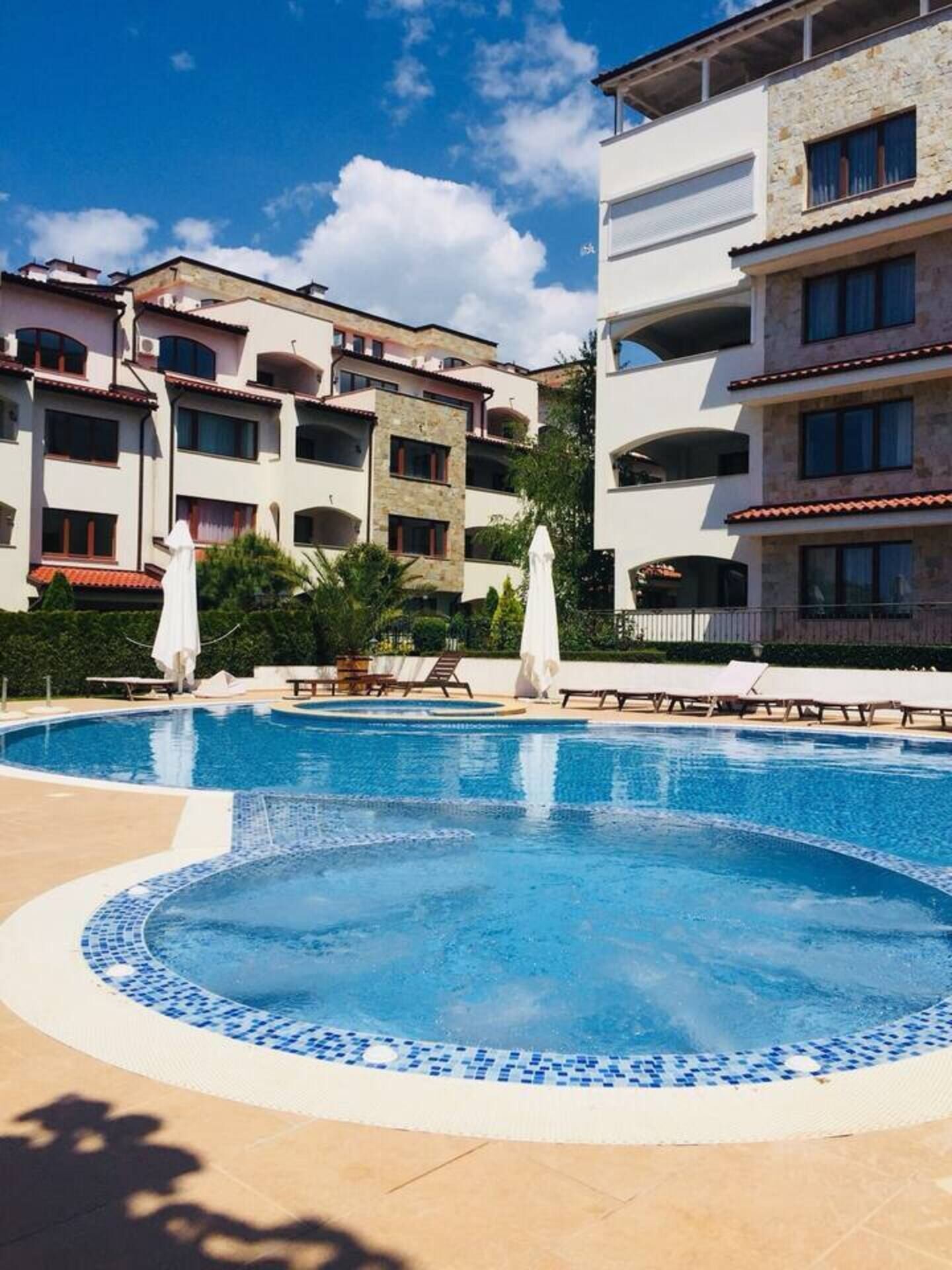 A swimming pool with lounge chairs and umbrellas, surrounded by multi-story buildings with balconies.