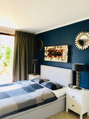 A bedroom with a bed, two bedside tables, a wall clock, a decorative wall piece, and a window with curtains.