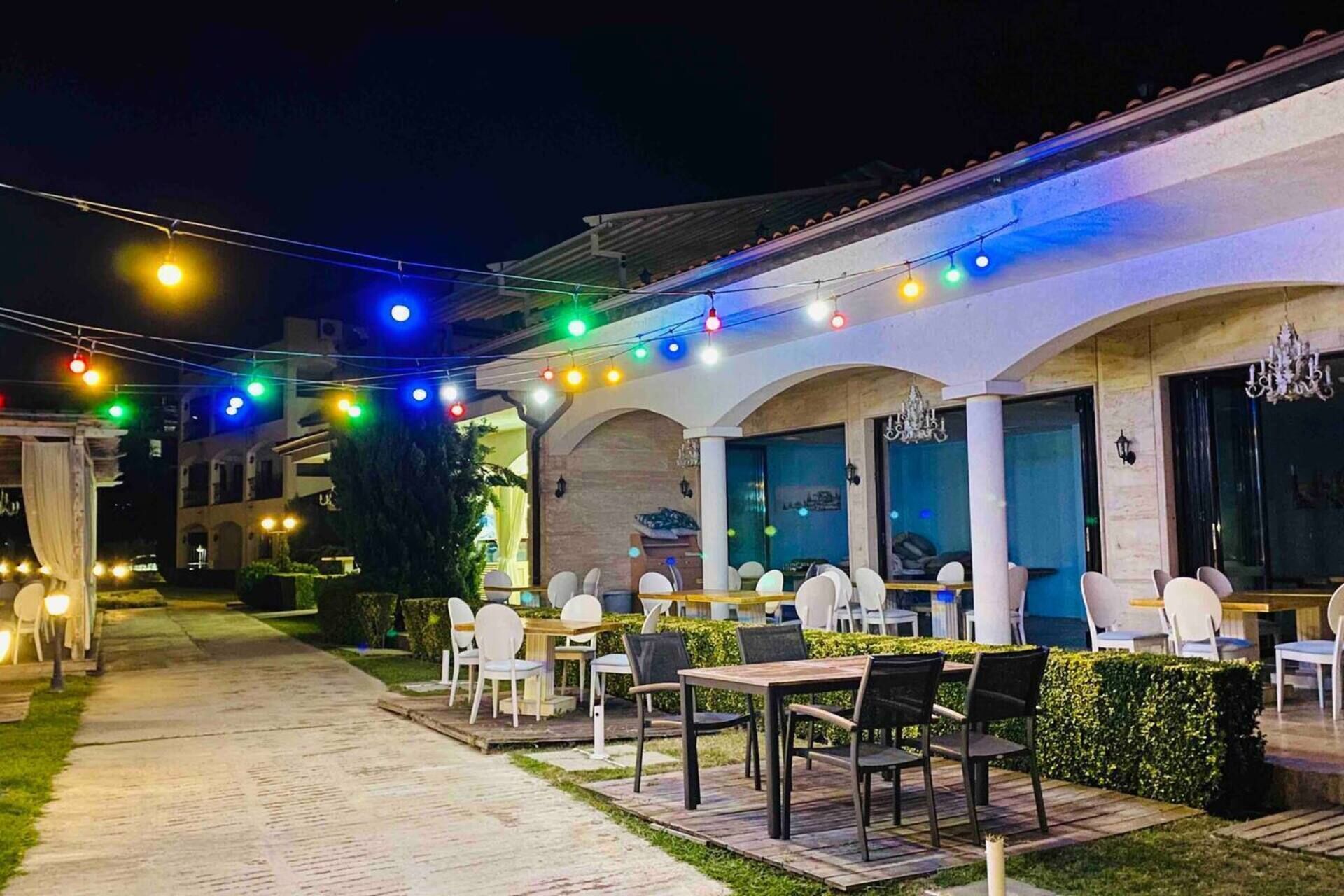 An outdoor dining area with tables and chairs, string lights overhead, and a pathway leading to the entrance.