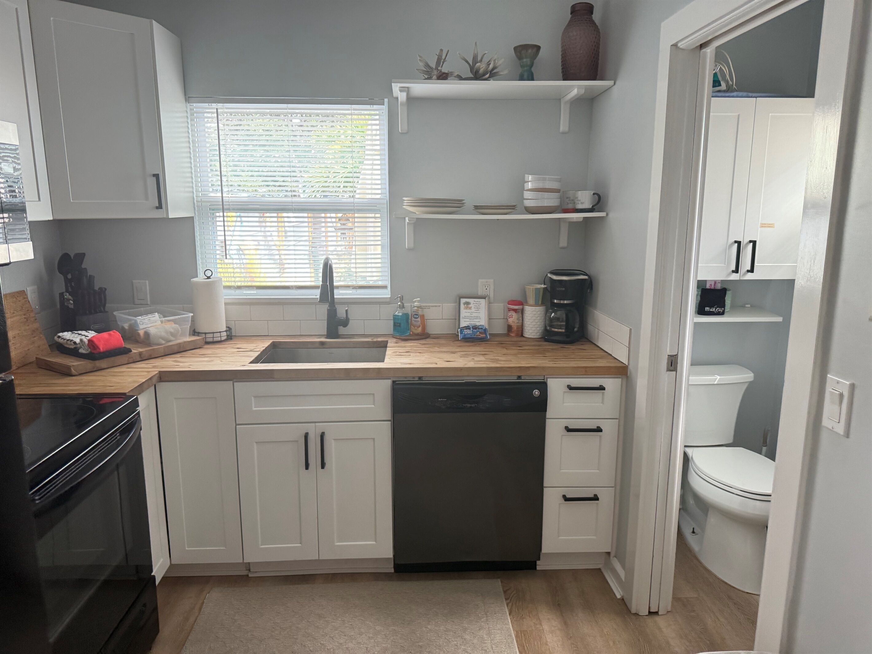 Una cocina moderna con armarios blancos, lavavajillas, cafetera y ventana con persianas.