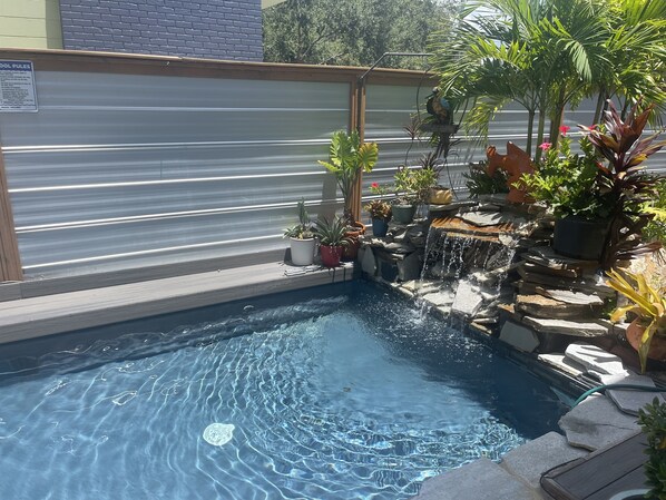 A swimming pool with a waterfall feature, surrounded by various potted plants and a metal fence.