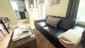 Una sala de estar con un sofá de cuero negro, una mesa de centro tejida y una cocina al fondo.