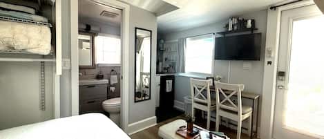 Una habitación de hotel compacta con cama, un escritorio pequeño, una silla, televisor, baño y una estantería con libros.