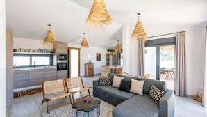Een moderne woonkamer met een grijze modulaire bank, rieten stoelen en een salontafel. Er zijn drie hangende rieten lampen, een houten kast en een eetgedeelte met een tafel en stoelen zichtbaar door een glazen deur.