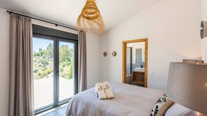 Een slaapkamer met een groot bed, een raam met gordijnen, een hanglamp en een spiegel aan de muur.